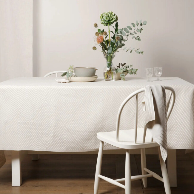 Gänseaugen-Tischdecke in der Farbe Weiß, auf einem Tisch ausgelegt. Ein Herrenhandtuch aus Asche hängt an einem Stuhl. 